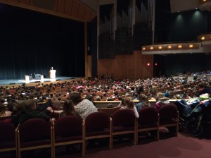 An auditorium photographed from the back. The seats are all filled. It is hard to make out, but R.L. Stine is standing at a podium on stage addressing the audience.