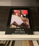 Image of a book laid on a display shelf in my bookcase. The book cover text: Voices of Jane Adeny Memorial School. Book cover picture is looking over the shoulder of a young Kenyan student as she writes.